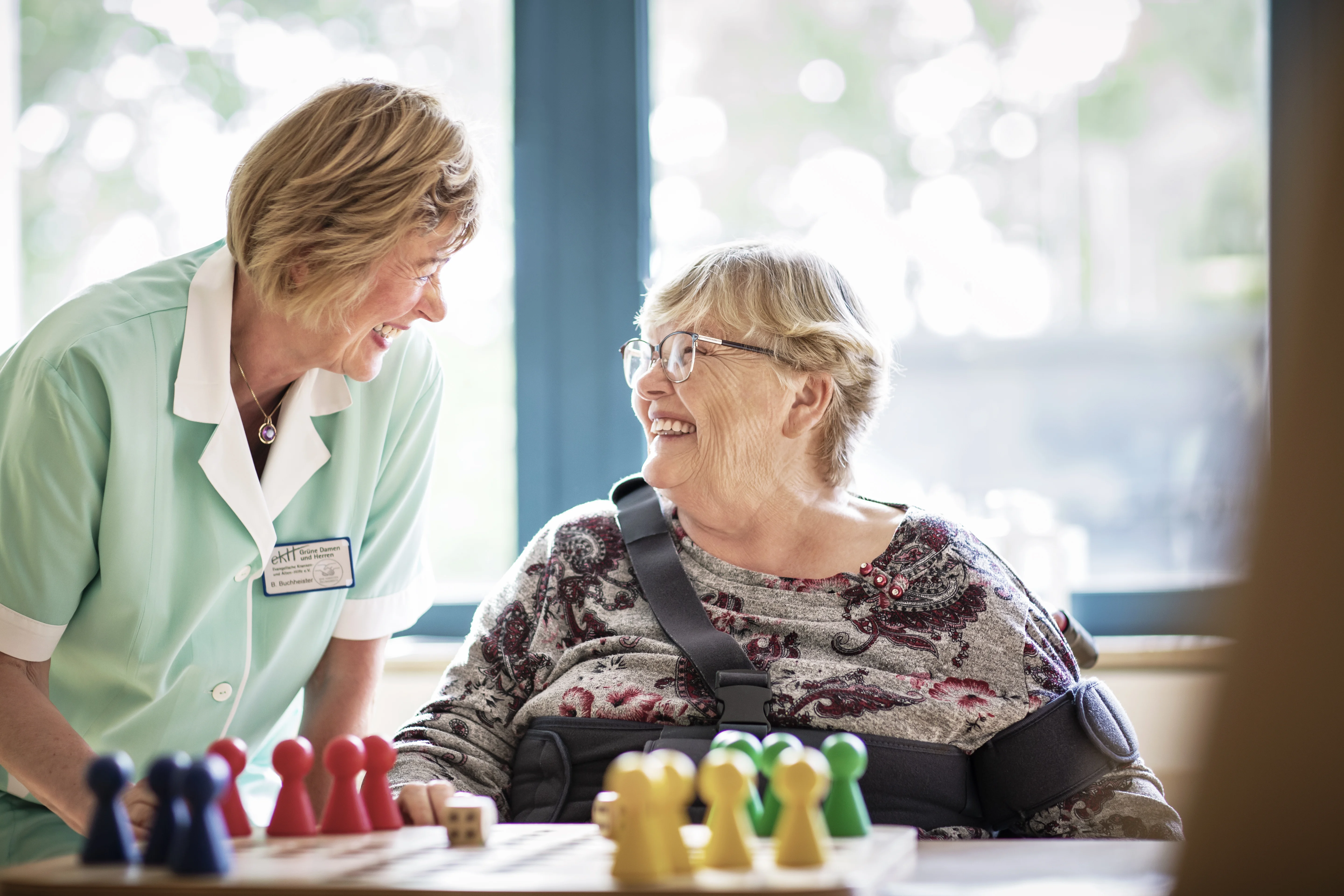 Eine ehrenamtliche Mitarbeiterin und eine Patientin. Beide lächeln sich sympathisch an. Im Vordergrund ein Brettspiel.