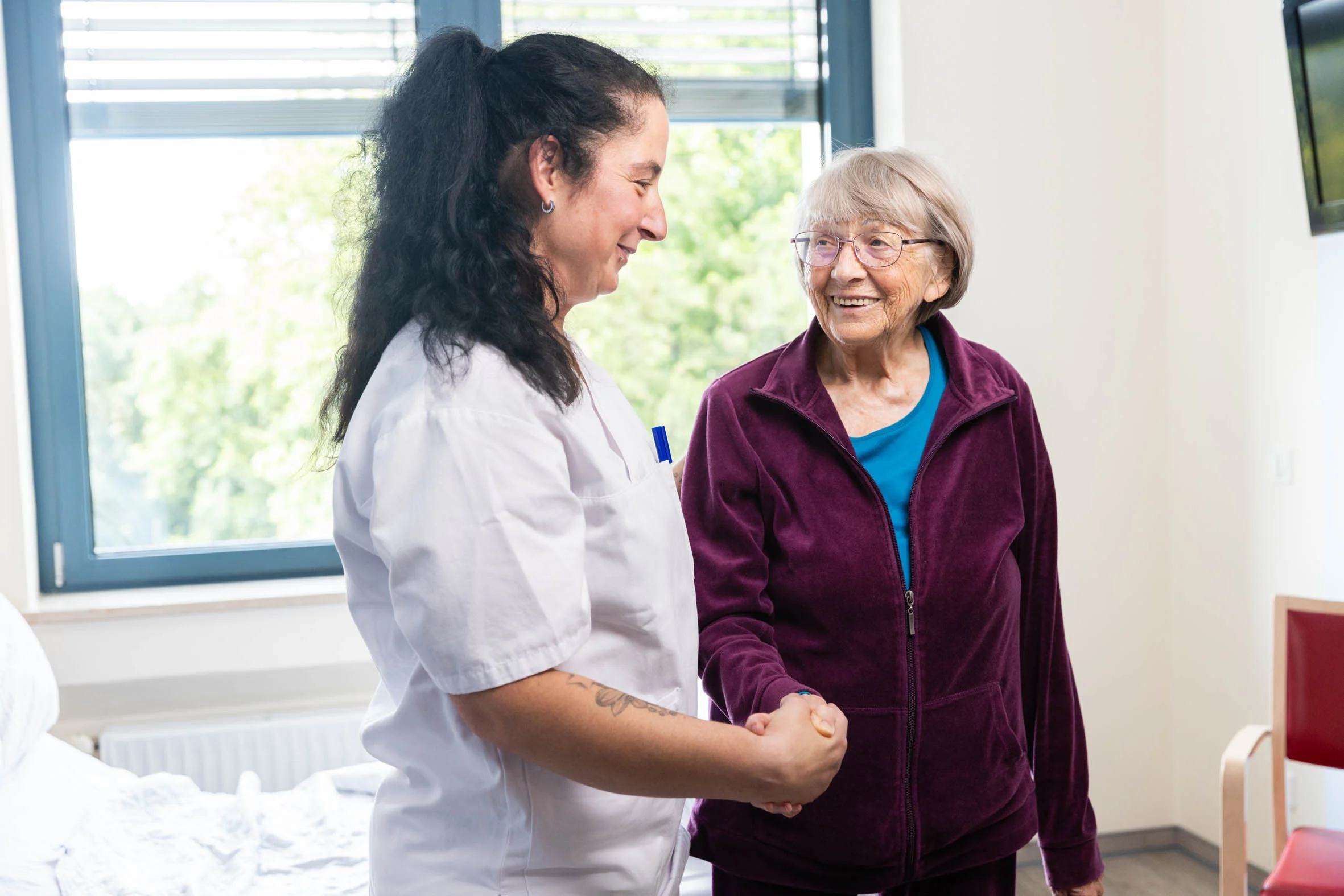 DianaKlinik | Patientenzufriedenheit Eine Pflegekraft und eine Patientin geben einander die Hände. Beide lächeln sich an.