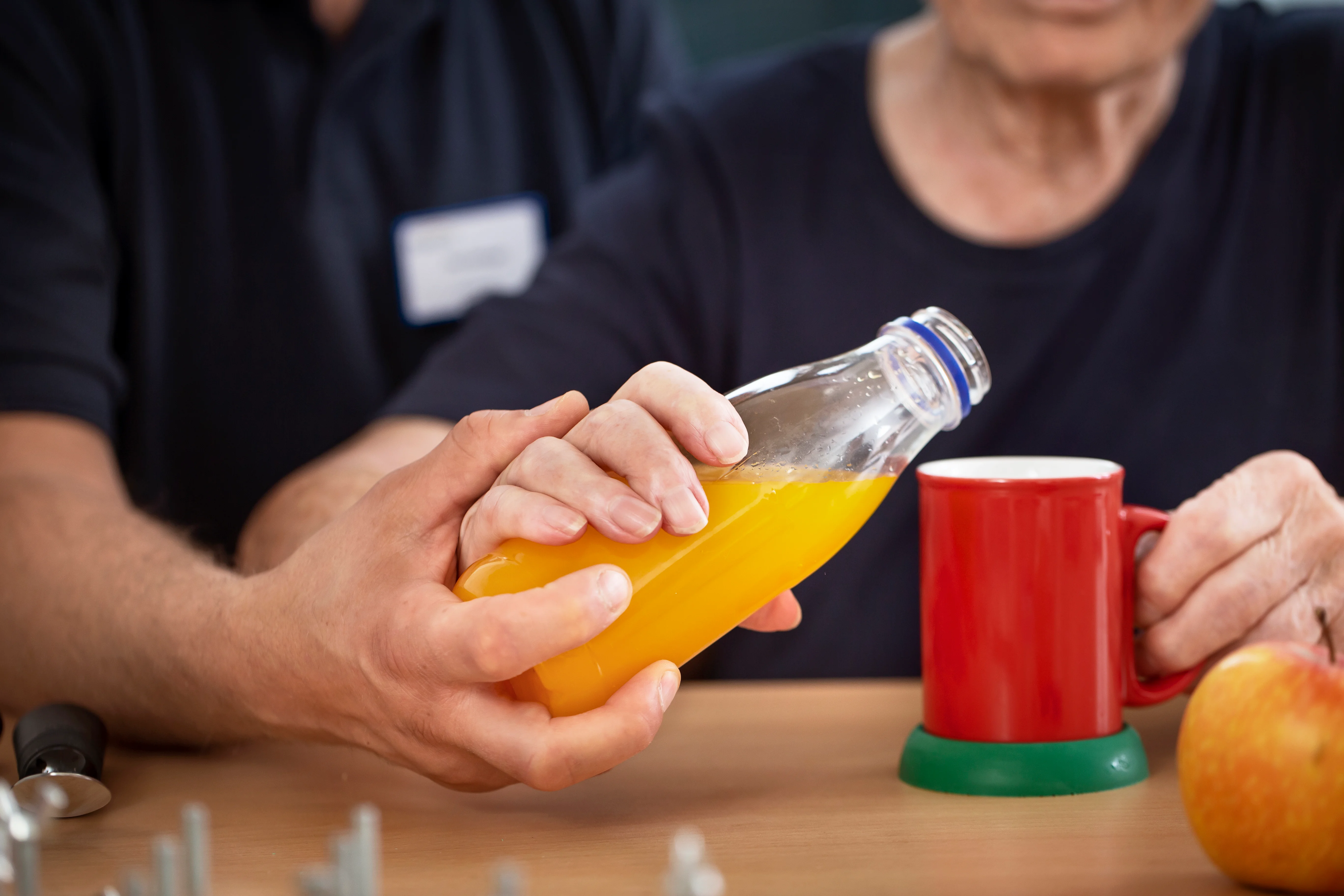 Eine Nahaufnahme. Eine Pflegekraft unterstützt eine Patientin bei der Motorik. Sie gießt ein Getränk in eine Tasse. 