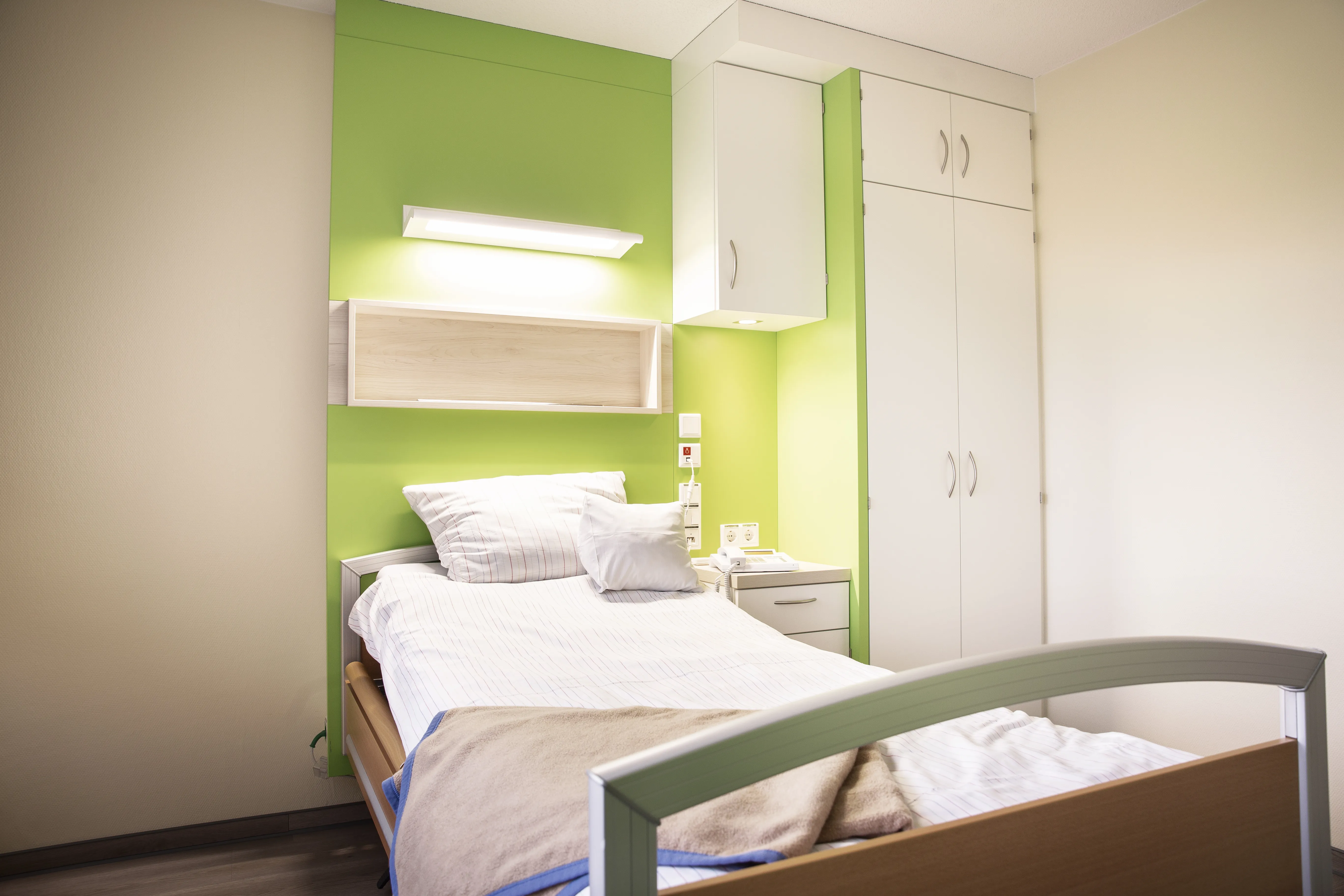 Die Möbel wie Bett und Schrank im Patientenzimmer sind aus hellem Holz mit akzentuierten, hellgrünen Wandelementen.