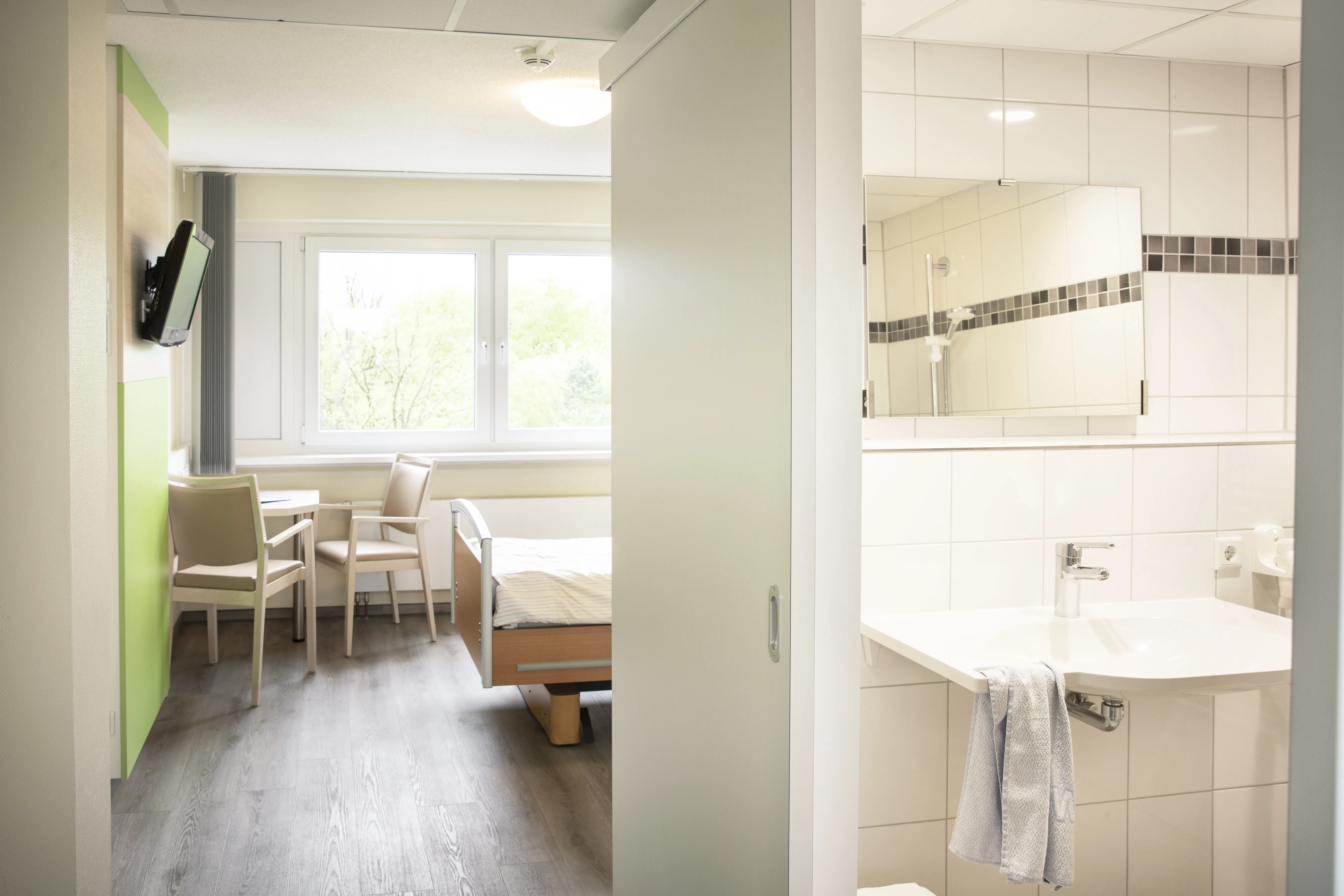 Blick in ein lichtdurchflutetes Patientenzimmer mit hellen Möbeln und weiß gefließtem Bad.
